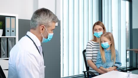 Caucasian-blonde-female-patient-sitting-in-hospital-cabinet-with-her-daughter-and-listening-to-senior-doctor-in-medical-mask,-general-practitioner-showing-vaccines-to-woman-on-consultation-virus-cure